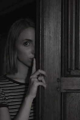 A person stands partially obscured by a wooden door, holding a finger to their lips in a gesture suggesting silence or secrecy. The lighting is dim, creating a mysterious and slightly ominous atmosphere.
