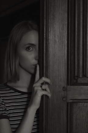 A person stands partially obscured by a wooden door, holding a finger to their lips in a gesture suggesting silence or secrecy. The lighting is dim, creating a mysterious and slightly ominous atmosphere.