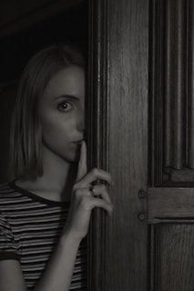 A person stands partially obscured by a wooden door, holding a finger to their lips in a gesture suggesting silence or secrecy. The lighting is dim, creating a mysterious and slightly ominous atmosphere.