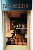 A cozy brasserie with an inviting interior characterized by warm lighting, wooden tables, and black chairs. The tiled floor features a geometric pattern in shades of brown and white. Mirrors and decorations adorn the walls, adding to the elegant and relaxed atmosphere.