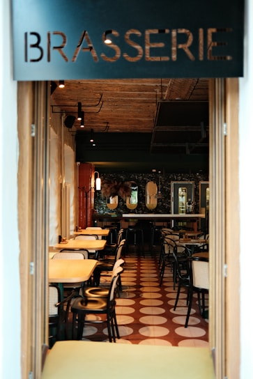 A cozy brasserie with an inviting interior characterized by warm lighting, wooden tables, and black chairs. The tiled floor features a geometric pattern in shades of brown and white. Mirrors and decorations adorn the walls, adding to the elegant and relaxed atmosphere.