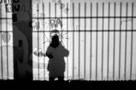The image features a shadow cast on a wall with graffiti. The graffiti includes the word 'GUERRA' and a depiction of a figure with a covered face and swords. Shadows from vertical bars are also visible, contributing to a sense of confinement.