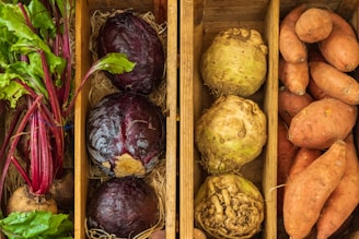 A wooden box is divided into sections containing fresh vegetables. On the left, there are beetroots with green leaves attached. The middle section contains two large red cabbages and two celeriac roots. The right section is filled with several sweet potatoes. All the vegetables are placed on a bed of straw.