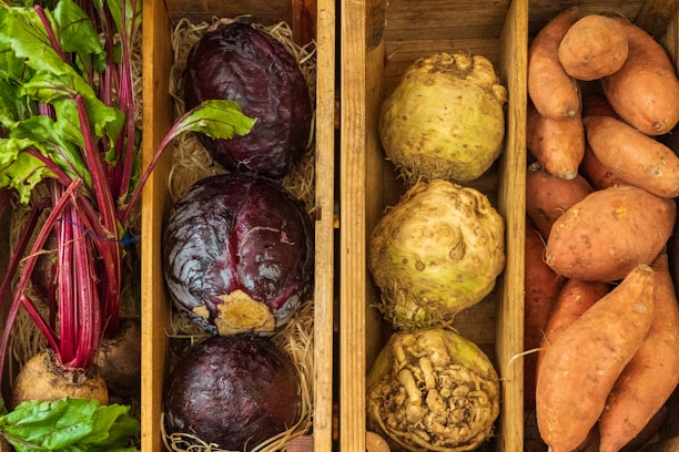 A wooden box is divided into sections containing fresh vegetables. On the left, there are beetroots with green leaves attached. The middle section contains two large red cabbages and two celeriac roots. The right section is filled with several sweet potatoes. All the vegetables are placed on a bed of straw.