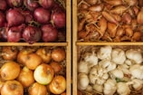 An arrangement of vegetables in four wooden crates positioned in a grid. The top left crate contains red onions, the top right has shallots, the bottom left holds yellow onions, and the bottom right is filled with bulbs of garlic. The crates are lined with straw-like material, providing a rustic feel.