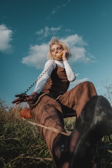 A person with blonde hair speaks into a yellow vintage telephone while sitting on grass. They are dressed in a brown outfit with a white long-sleeve shirt. The scene appears to be in a field under a blue sky with scattered clouds.