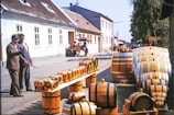 A street scene featuring two men in suits, standing and inspecting a display of wooden barrels and small casks arranged on wooden planks. The barrels are various sizes and are set along the roadside in front of a row of houses. Behind the men, there is a group of people under a red umbrella, engaged in activities possibly at a market or festival. The architecture features white buildings with tiled roofs, and the day appears to be bright and sunny.