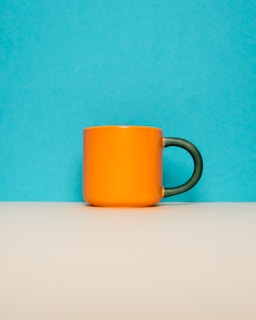 An orange mug with a dark green handle is placed on a beige surface against a bright blue background. The composition is simple and minimalist.