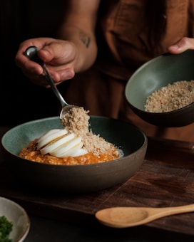 A person is preparing a dish by sprinkling a crumbly substance over a bowl that contains a sliced boiled egg on top of what appears to be a cooked grain or porridge. The scene suggests a focus on culinary preparation, with hands carefully adding ingredients. The setting includes a wooden surface with a wooden spoon nearby, creating a rustic and homely ambiance.