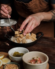 A person is preparing a dish by placing cubes of tofu on top of a bed of rice. The person is wearing a brown apron and holding a spoon in one hand while arranging the tofu with the other. On the table, there are small bowls containing sliced jalapeños, crushed peanuts, and a diced salsa mixture, along with lemon wedges.