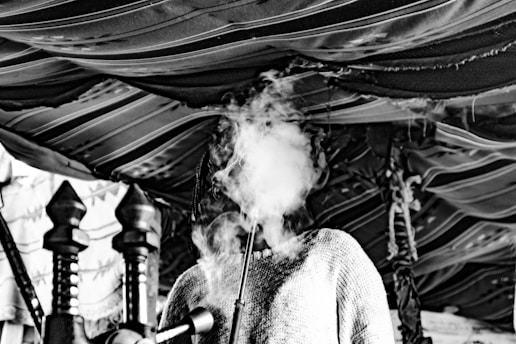 A person with a head full of smoke, obscuring their face, stands under a fabric canopy adorned with wooden poles. The setting appears to be indoors, with patterns and textures highlighting a rustic environment.
