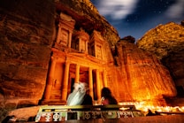 A magnificent rock-cut architecture illuminated at night, showcasing intricate carvings and columns. The scene is enhanced by flickering candle lights lining the base, with a clear starry sky overhead. Two figures are seated in front of this ancient structure, facing it and possibly enjoying the serene atmosphere.