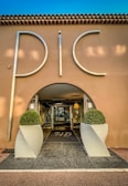 An elegant entrance to a building with large silver letters spelling 'PIC', flanked by two symmetrical potted topiary arrangements in white, modern planters. The structure has a stucco facade with a tiled roof, and the interior visible through a glass door is warmly lit.