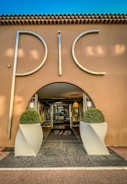 An elegant entrance to a building with large silver letters spelling 'PIC', flanked by two symmetrical potted topiary arrangements in white, modern planters. The structure has a stucco facade with a tiled roof, and the interior visible through a glass door is warmly lit.