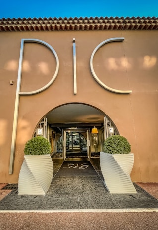 An elegant entrance to a building with large silver letters spelling 'PIC', flanked by two symmetrical potted topiary arrangements in white, modern planters. The structure has a stucco facade with a tiled roof, and the interior visible through a glass door is warmly lit.