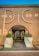 An elegant entrance to a building with large silver letters spelling 'PIC', flanked by two symmetrical potted topiary arrangements in white, modern planters. The structure has a stucco facade with a tiled roof, and the interior visible through a glass door is warmly lit.