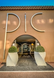 An elegant entrance to a building with large silver letters spelling 'PIC', flanked by two symmetrical potted topiary arrangements in white, modern planters. The structure has a stucco facade with a tiled roof, and the interior visible through a glass door is warmly lit.
