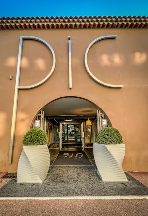 An elegant entrance to a building with large silver letters spelling 'PIC', flanked by two symmetrical potted topiary arrangements in white, modern planters. The structure has a stucco facade with a tiled roof, and the interior visible through a glass door is warmly lit.