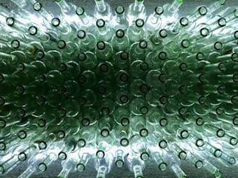 A collection of clear glass bottles arranged in a symmetrical pattern, viewed from above. The bottles are illuminated from below, creating a glowing effect, with the green tint of the glass adding a unique texture to the image.