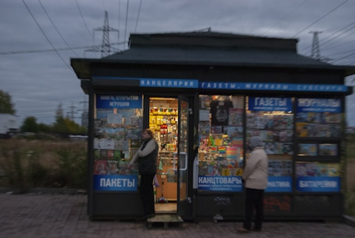 A small kiosk or newsstand with various magazines, newspapers, and stationery items displayed in the windows. Two people are standing near the entrance, one looking at the displays.