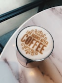 A cup of coffee on a marble table, with a decorative foam design that reads 'Life begins after coffee'. The design includes ornate, swirling patterns above and below the text.