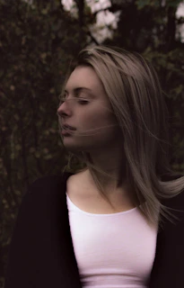 A woman with long hair is seen in profile with her eyes closed, surrounded by a dark and blurred natural background. She wears a light-colored top and a dark outer garment. The atmosphere suggests a moment of quiet reflection in a secluded outdoor setting.