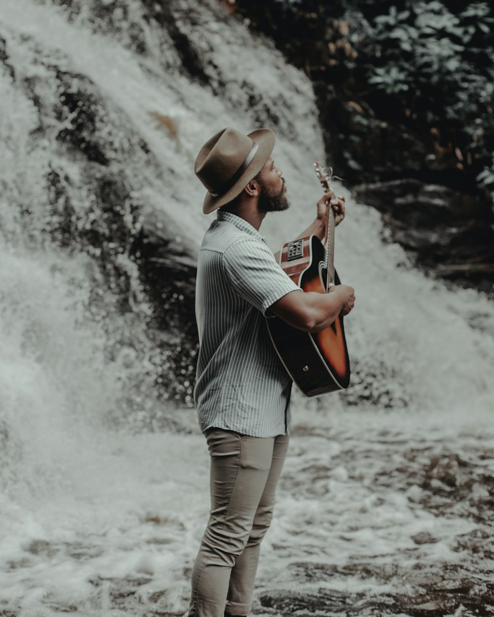 a man holding a guitar standing in front of a waterfall