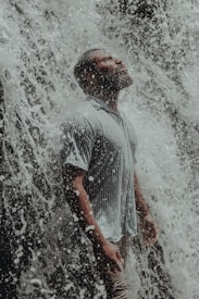 A person stands with eyes closed and head tilted slightly upward, surrounded by a cascade of water from a waterfall. Their face and clothing are wet, creating a serene and contemplative atmosphere.