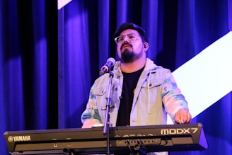 A musician is playing a Yamaha MODX7 keyboard on stage. They are wearing glasses and a colorful tie-dye jacket. A microphone stand is positioned in front of them, and blue stage curtains serve as the backdrop, accentuated by bright stage lighting.