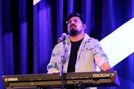 A musician is playing a Yamaha MODX7 keyboard on stage. They are wearing glasses and a colorful tie-dye jacket. A microphone stand is positioned in front of them, and blue stage curtains serve as the backdrop, accentuated by bright stage lighting.