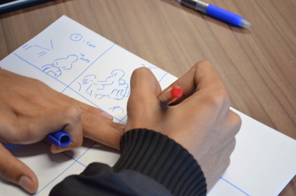 A person is sketching a storyboard on a piece of paper, using blue ink. The storyboard contains simplified drawings, likely for a visual or media project, and is held on a wooden table surface. A pen with a red cap is held while another hand holds a blue pen. Nearby on the table, there is another pen with a blue cap.