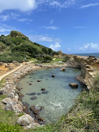 A scenic coastline features clear turquoise water bordered by rocky formations and lush greenery. A dirt path runs adjacent to the shore, leading to distant hills covered with dense vegetation. The sky above is bright with scattered clouds.