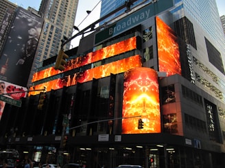 A bustling cityscape with a modern building adorned with vibrant, illuminated digital screens displaying fiery, abstract designs. The surroundings feature towering skyscrapers and busy traffic lights. Pedestrians and vehicles are present, adding to the urban atmosphere. The backdrop includes large billboards and street signs.
