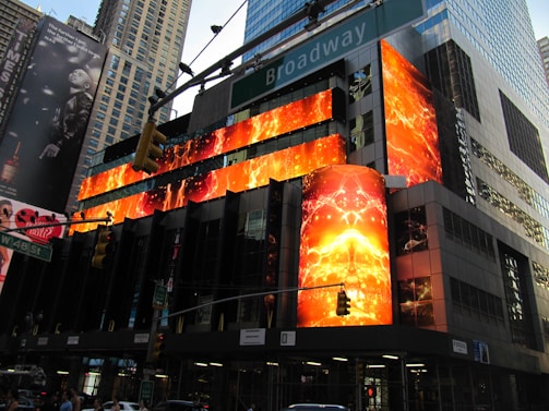 A bustling cityscape with a modern building adorned with vibrant, illuminated digital screens displaying fiery, abstract designs. The surroundings feature towering skyscrapers and busy traffic lights. Pedestrians and vehicles are present, adding to the urban atmosphere. The backdrop includes large billboards and street signs.