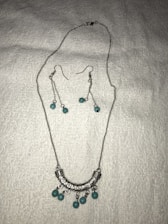A set of jewelry featuring a silver necklace and matching earrings is displayed on a white textured background. The necklace has a curved, engraved silver pendant adorned with turquoise beads. The earrings feature long silver chains with matching turquoise beads.