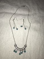 A set of jewelry featuring a silver necklace and matching earrings is displayed on a white textured background. The necklace has a curved, engraved silver pendant adorned with turquoise beads. The earrings feature long silver chains with matching turquoise beads.