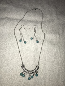 A set of jewelry featuring a silver necklace and matching earrings is displayed on a white textured background. The necklace has a curved, engraved silver pendant adorned with turquoise beads. The earrings feature long silver chains with matching turquoise beads.