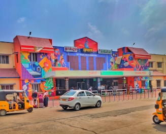 A brightly colored train station building featuring vivid murals with various characters and patterns. Several people are gathered near the entrance, and parked vehicles, including an auto rickshaw and a car, are visible in the foreground. Signage in multiple languages is displayed, indicating it is the Tambaram station.