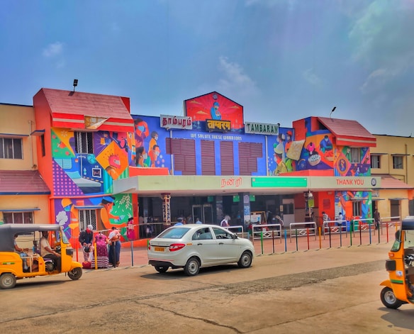 A brightly colored train station building featuring vivid murals with various characters and patterns. Several people are gathered near the entrance, and parked vehicles, including an auto rickshaw and a car, are visible in the foreground. Signage in multiple languages is displayed, indicating it is the Tambaram station.