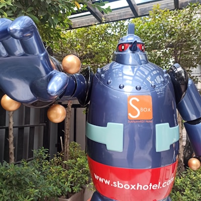 A large, blue robot statue with a shiny surface is positioned outdoors. It has a red band with a logo and website URL on its torso. The robot is surrounded by green plants and a glass canopy overhead.