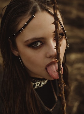 A young person with dark eye makeup and hair adorned with metal rings is depicted in an edgy and unconventional pose, licking a thorny branch. The background is a blurred natural setting, adding a rustic and possibly autumnal feel to the composition.