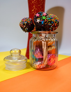 A glass jar filled with colorful candy and topped with chocolate-covered cake pops decorated with vibrant sprinkles and star-shaped toppings. The jar is placed next to a detached glass lid and is set against a bright yellow and orange surface with a red decorative object in the background.