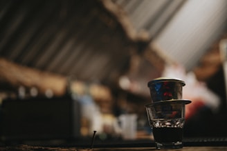 In a dimly lit setting, a traditional Vietnamese coffee drip is placed atop a glass filled with dark coffee. The background is blurred, highlighting the rustic environment.