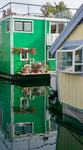 A vibrant green houseboat with a rooftop patio and potted plants is reflected in the calm water. Adjacent is a yellow houseboat with a small window showcasing a cozy interior with flowers. The peaceful setting highlights the unique lifestyle on the water.