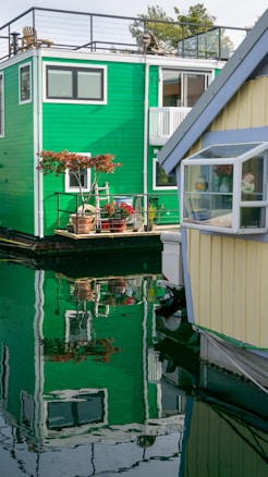 A vibrant green houseboat with a rooftop patio and potted plants is reflected in the calm water. Adjacent is a yellow houseboat with a small window showcasing a cozy interior with flowers. The peaceful setting highlights the unique lifestyle on the water.