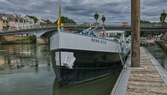A boat named Nanajesa is docked along a wooden pier on a calm river, with a bridge in the background. The cloudy sky adds a moody feel, and small European-style buildings line the riverbank, contributing to a picturesque cityscape.