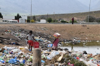 Two children are standing in a field filled with garbage, surrounded by litter and debris. One child carries a red container while the other balances a cardboard box on their head. In the background, a road, several vehicles, and mountains can be seen under a cloudy sky.