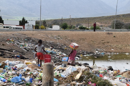 Two children are standing in a field filled with garbage, surrounded by litter and debris. One child carries a red container while the other balances a cardboard box on their head. In the background, a road, several vehicles, and mountains can be seen under a cloudy sky.