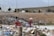 Two children are standing in a field filled with garbage, surrounded by litter and debris. One child carries a red container while the other balances a cardboard box on their head. In the background, a road, several vehicles, and mountains can be seen under a cloudy sky.