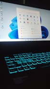 A laptop with a glowing backlit keyboard and a desktop screen displaying various icons and applications. The keyboard emits a blue-green light, creating a futuristic look. The desktop screen shows a start menu with pinned and recommended applications, set against a light blue background.